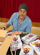 Charger l'image dans la galerie, Artist Jonathon McDaniel wearing a gray beanie and a light blue button-up shirt sits behind a table covered with printed comic book or graphic novel copies featuring Dracula. An open cardboard box is on the left, and a red curtain hangs in the background.